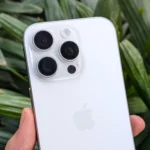 iPhone 16 Pro màu trắng được cầm trên tay trước nền cây xanh, minh họa thiết kế tổng thể của camera.