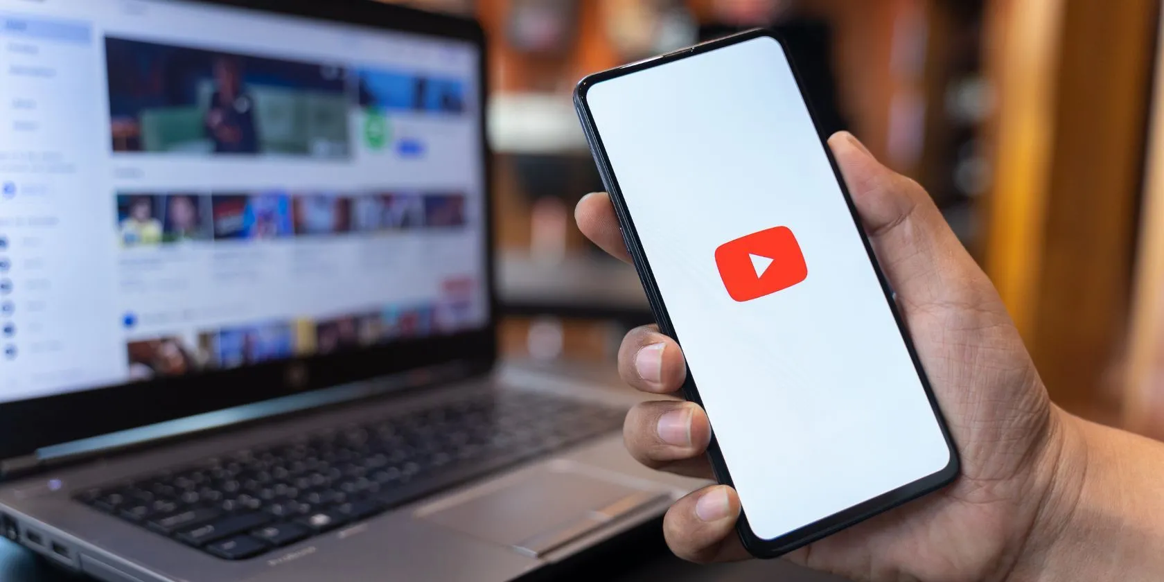 Người dùng đang cầm điện thoại hiển thị ứng dụng YouTube, với giao diện trang web YouTube phía sau