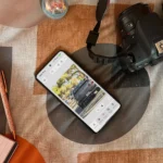 Người dùng đang sử dụng ứng dụng Photoshop trên điện thoại Android đặt trên giường, bên cạnh là máy ảnh và sổ tay, thể hiện sự tiện lợi khi làm việc di động.